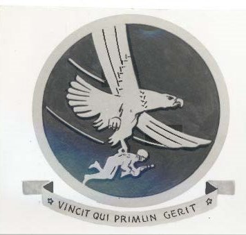 A grayscale emblem of the Troop Carrier Command depicting a stylized condor clutching in its dexter claw a Paratrooper carrying a "tommy" gun, ready for action. On the scroll: Vincit Qui Primum Gerit (He conquers who gets there first).